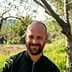 Man with a beard smiling while standing between olive trees in a natural setting. - Olive Oil Times