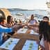 A group of five people toasting with drinks at a wooden table overlooking a coastal view. - Olive Oil Times