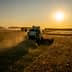 A harvesting machine working in a field during sunset, with dust rising in the air. - Olive Oil Times