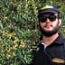 Man wearing sunglasses and a black cap standing next to an olive tree with green olives. - Olive Oil Times