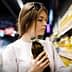 Woman with brown hair holding a bottle of olive oil while examining products on a shelf in a grocery store. - Olive Oil Times