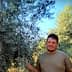 A man standing next to olive trees in an outdoor setting, with green foliage in the background. - Olive Oil Times