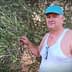 Man in a tank top and blue cap standing next to an olive tree while holding a branch. - Olive Oil Times
