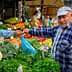 A man in a blue cap interacts with a vendor at a vegetable market filled with various fresh produce. - Olive Oil Times