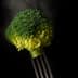 A piece of steamed broccoli held by a fork against a dark background with steam rising. - Olive Oil Times