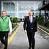 Three men walking on a pathway, two wearing green vests and one in a suit. - Olive Oil Times