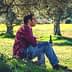 A man in a red plaid shirt sitting on a rock under an olive tree, holding a bottle. - Olive Oil Times