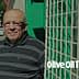 Man wearing glasses and a striped shirt standing next to a green fence with a grid pattern. - Olive Oil Times