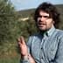 Man wearing a checkered shirt gesturing while standing in an olive grove. - Olive Oil Times