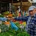 Man in a blue plaid shirt interacting with a vendor at a vegetable market stall. - Olive Oil Times