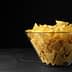 A clear glass bowl filled with yellow tortilla chips on a dark surface. - Olive Oil Times