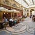 People dining at tables in a covered arcade with a glass roof and decorative floor tiles. - Olive Oil Times