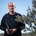 Man standing outdoors holding a bottle of olive oil next to an olive tree. - Olive Oil Times