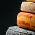 A stack of different types of cheese, including a yellow-orange cheese and a dark cheese, against a dark background. - Olive Oil Times
