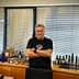 Man with gray hair wearing a black t-shirt standing in an office with olive oil products displayed on a shelf. - Olive Oil Times