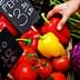 A hand reaching for red and yellow bell peppers among various vegetables at a market stand. - Olive Oil Times