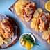 Four lobster rolls served with potato chips and lemon slices on a wooden platter. - Olive Oil Times
