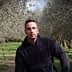 A man wearing a black hoodie sitting among olive trees in an olive grove. - Olive Oil Times