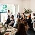A speaker presenting information on a screen to an audience seated at a long table during a meeting. - Olive Oil Times