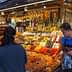 A market stall displaying various dried fruits, nuts, and bottles of olive oil with a vendor assisting a customer. - Olive Oil Times