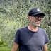 Man wearing a black cap and glasses smiling while standing in an olive grove. - Olive Oil Times