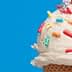 Close-up of an ice cream cone topped with vanilla ice cream and colorful sprinkles against a blue background. - Olive Oil Times