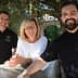Three people standing together outdoors, smiling and posing for a photo near an olive tree. - Olive Oil Times
