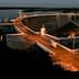 Concrete dam structure with a roadway and lights at dusk, reflecting in the water below. - Olive Oil Times