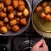 A bowl filled with golden-brown fried dough balls next to a frying pan with more dough balls being drained. - Olive Oil Times