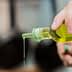 A person pouring olive oil from a glass bottle with a green cap, with oil dripping from the spout. - Olive Oil Times