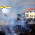 Yellow firefighting airplane releasing water over a wildfire near a white house. - Olive Oil Times