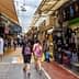 Two people walking through a market street lined with shops and stalls selling various items. - Olive Oil Times