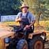 A man sitting on a yellow tractor wearing a straw hat and overalls, giving a thumbs up. - Olive Oil Times