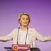 A woman gesturing while speaking at a podium during an event with a purple background. - Olive Oil Times