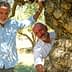 Two men standing beside a large olive tree, one leaning against the trunk and the other holding onto a branch. - Olive Oil Times