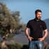 Man wearing a black polo shirt and jeans standing outdoors with olive trees in the background. - Olive Oil Times