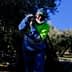 A worker in a green and blue outfit harvesting olives in an olive grove under a clear blue sky. - Olive Oil Times