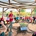 A group of children and adults seated in a circular outdoor learning space with a speaker in the center. - Olive Oil Times