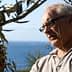 A man examining an olive tree branch while standing outdoors near the sea. - Olive Oil Times