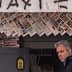 A man standing in front of a newsstand displaying various newspapers and magazines. - Olive Oil Times