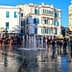 A fountain in a public square surrounded by buildings and people walking. - Olive Oil Times