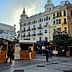 Wooden market stalls set up in an urban square with buildings in the background. - Olive Oil Times