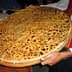 A person holding a large round flatbread topped with seeds and spices, presented on a wooden tray. - Olive Oil Times