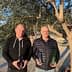 Two men standing outdoors holding bottles of olive oil in front of an olive tree. - Olive Oil Times