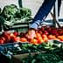 A hand reaching for ripe red tomatoes at a market stall filled with fresh vegetables. - Olive Oil Times