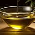 A clear glass bowl filled with olive oil, with olive branches placed nearby on a wooden surface. - Olive Oil Times