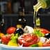 A bowl of fresh salad being drizzled with olive oil, featuring tomatoes, lettuce, and olives. - Olive Oil Times