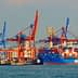 Large blue container ship docked at a port with red and blue cranes in the background. - Olive Oil Times