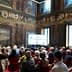 Audience seated in a historic hall during a conference presentation with decorative walls and a large screen. - Olive Oil Times