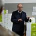 A man in a suit presenting a container of olive oil in a warehouse setting. - Olive Oil Times